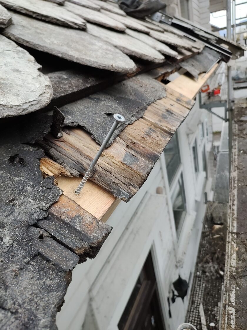 Uszkodzony dach z łupka z widocznym spróchniałym deskowaniem i wystającym wkrętem, ukazujący potrzebę wymiany dachu.