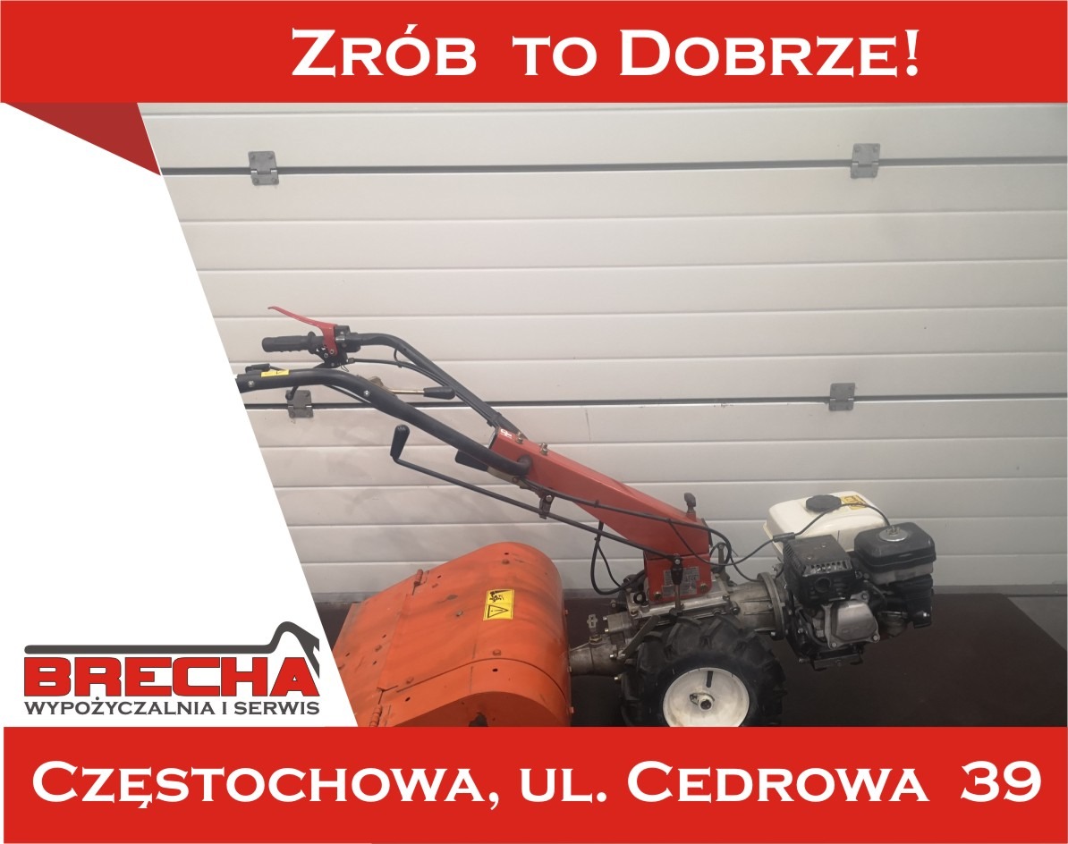 Pomarańczowy glebogryzarka spalinowa z białym zbiornikiem paliwa na tle białej bramy garażowej w pomieszczeniu.