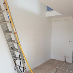 Marek Rulak - Wnętrze w trakcie prac wykończeniowych: białe ściany, aluminiowa drabina Krause z żółtymi elementami, pomarańczowe linie wyznaczające obszar roboczy na podłodze, widoczne ślady po remoncie.