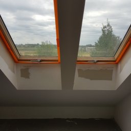 Marek Rulak - Dwa okna dachowe z widokiem na krajobraz, zabezpieczone pomarańczową taśmą malarską, ściany w trakcie przygotowania do malowania, widoczne ślady po szpachlowaniu.