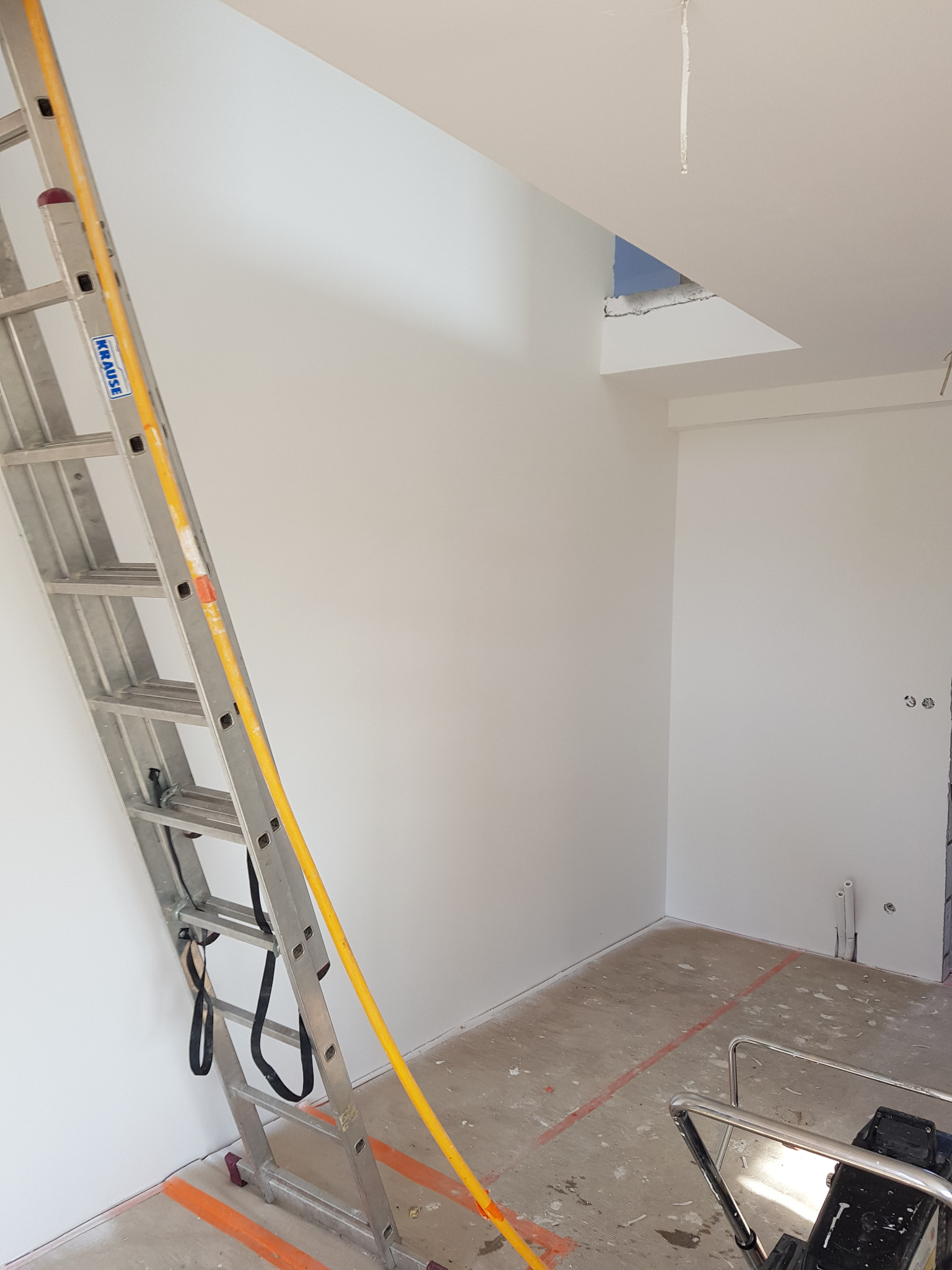 Wnętrze w trakcie prac wykończeniowych: białe ściany, aluminiowa drabina Krause z żółtymi elementami, pomarańczowe linie wyznaczające obszar roboczy na podłodze, widoczne ślady po remoncie.