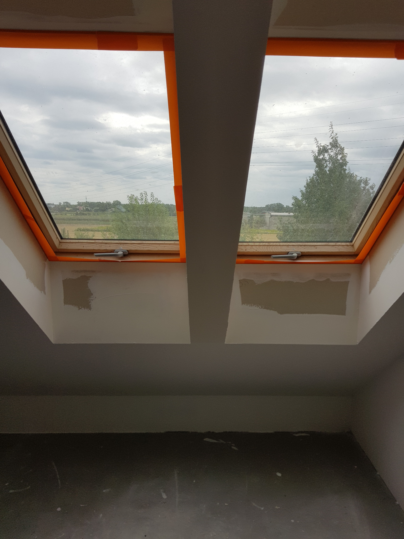 Dwa okna dachowe z widokiem na krajobraz, zabezpieczone pomarańczową taśmą malarską, ściany w trakcie przygotowania do malowania, widoczne ślady po szpachlowaniu.