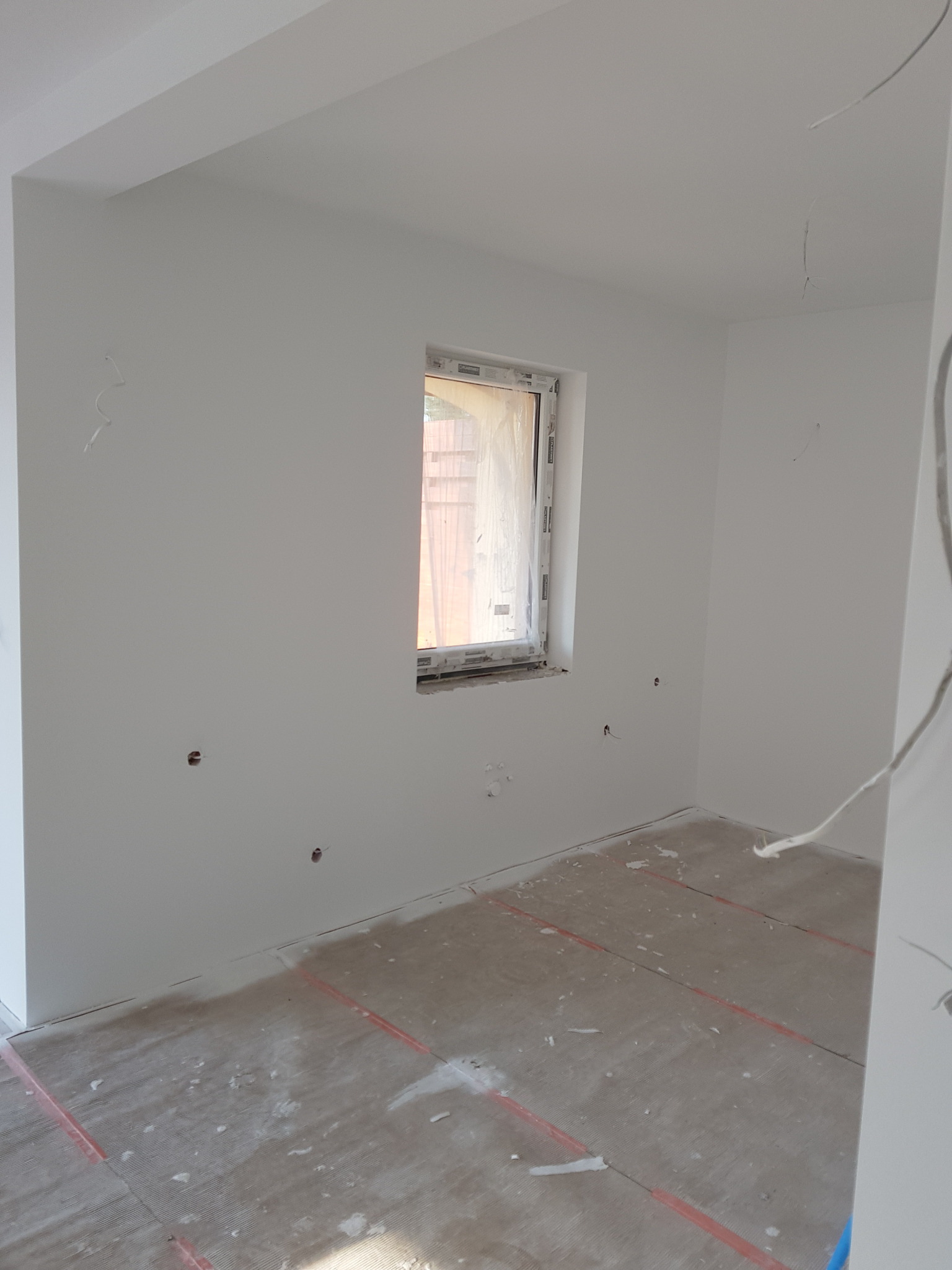 Wnętrze pokoju po wstępnym wyrównaniu ścian, z widocznymi otworami pod gniazdka elektryczne i zabezpieczonym oknem folią malarską.