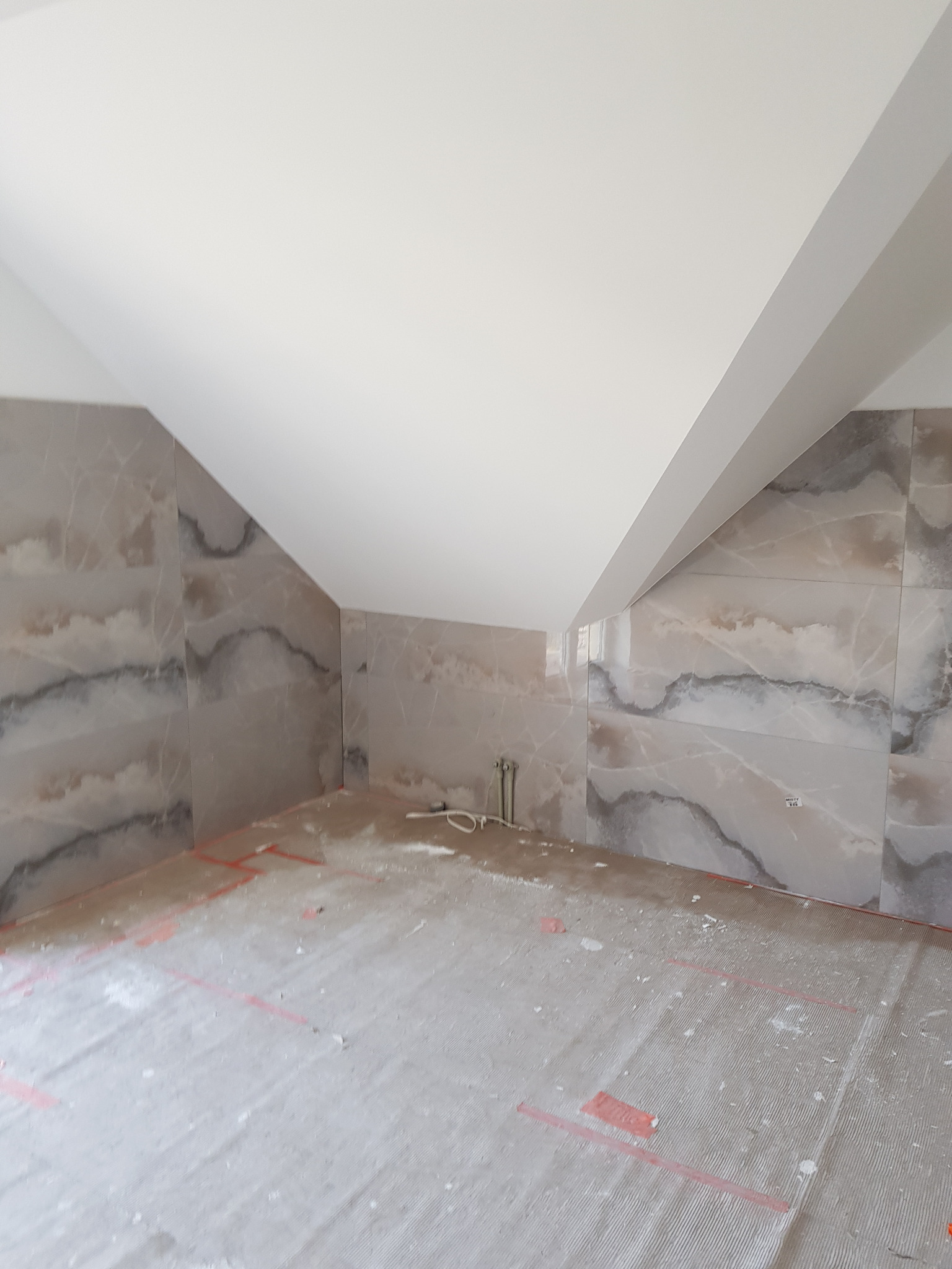 Poddasze w trakcie remontu, ściany wyłożone płytkami imitującymi marmur, sufit biały, podłoga przygotowana do dalszych prac wykończeniowych, widoczne ślady zaprawy.