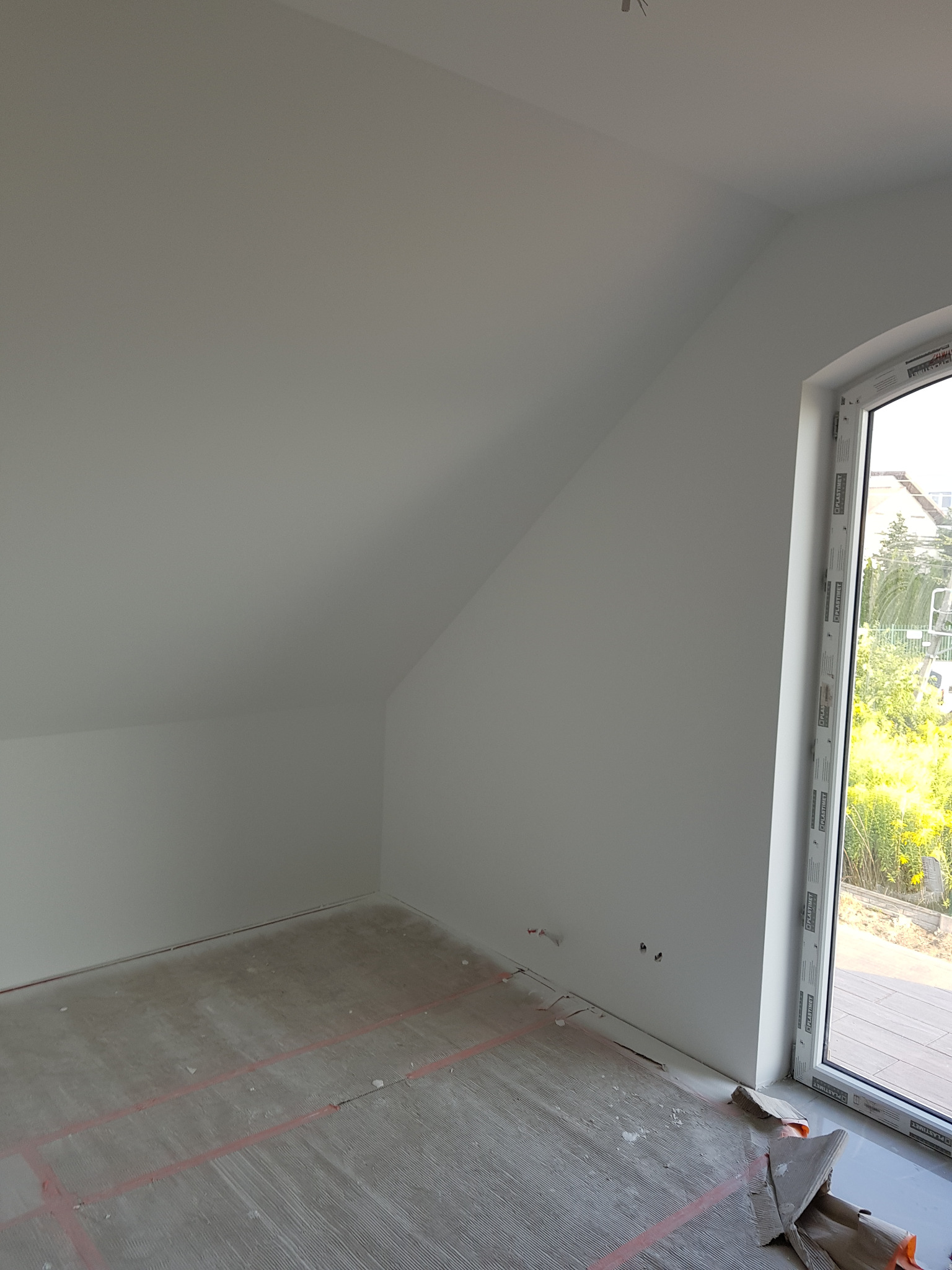 Poddasze po świeżym, białym wykończeniu ścian, widoczne przygotowane podłoże pod posadzkę z zaznaczonymi liniami oraz okno PCV z widokiem na zieleń.