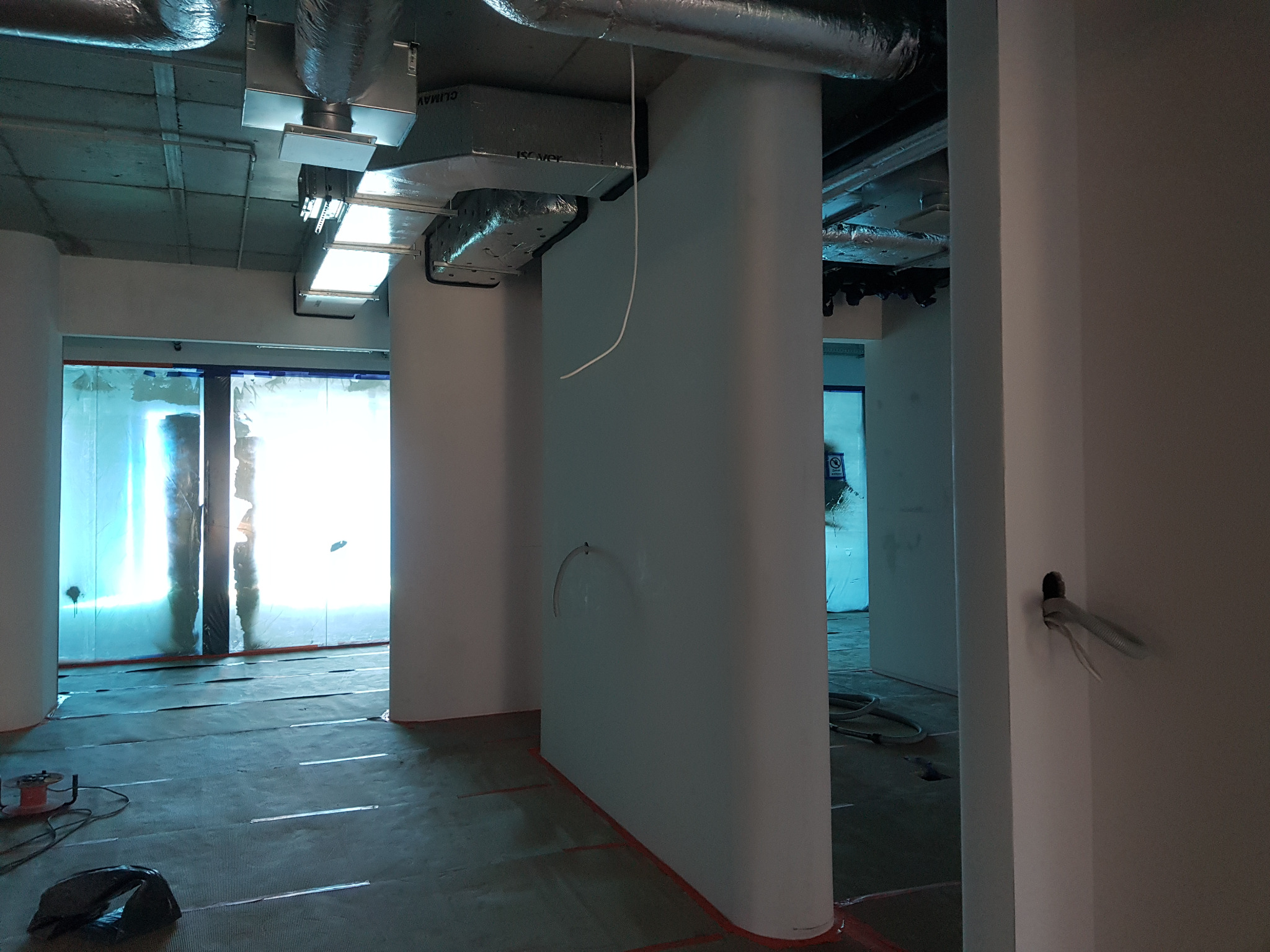 Wnętrze biurowe w trakcie prac wykończeniowych: zabezpieczone folią okna, przygotowana podłoga, białe, zaokrąglone kolumny, widoczne instalacje pod sufitem i przewody elektryczne wystające ze ściany.