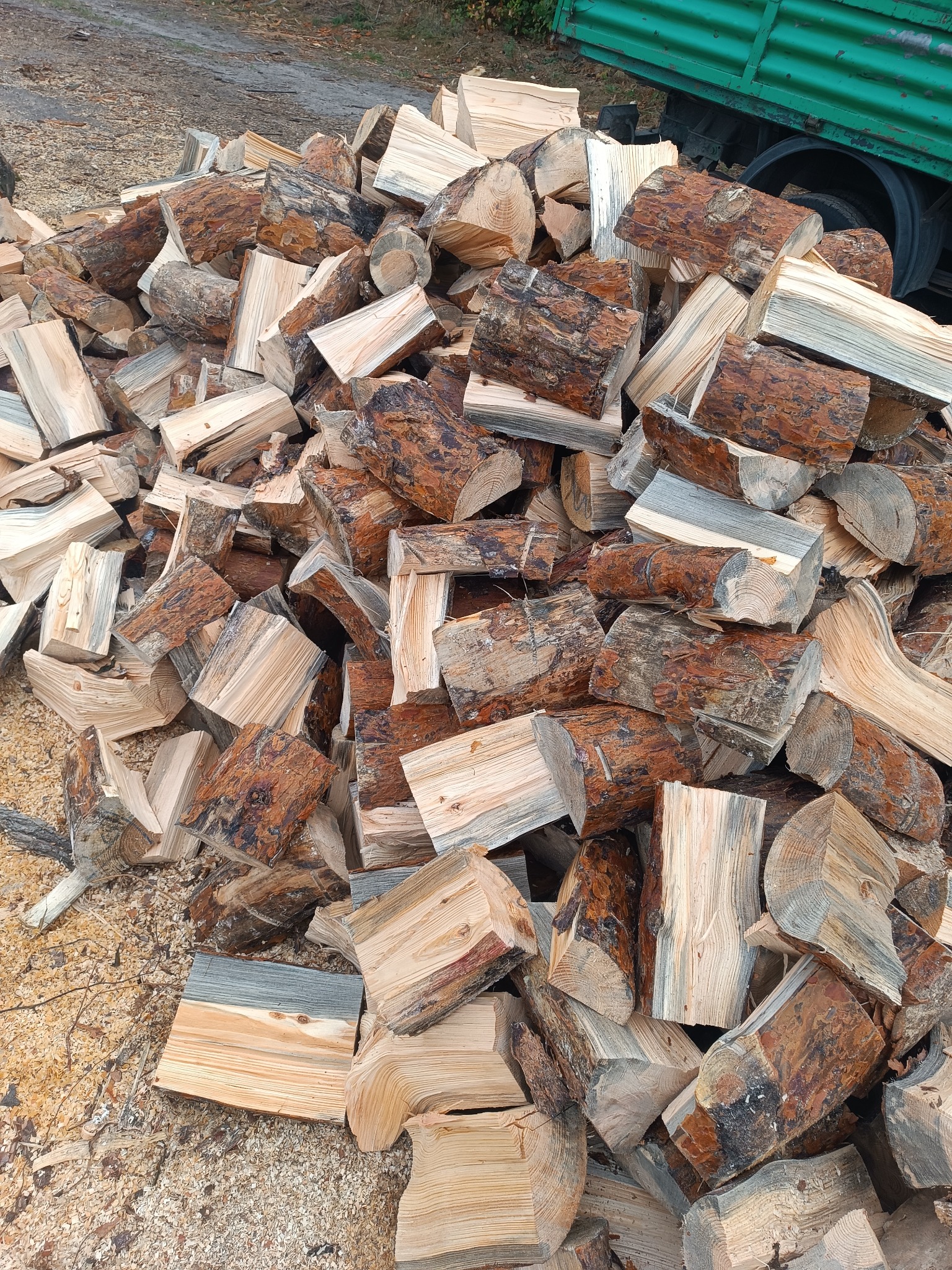 Stos pociętego drewna kominkowego z widoczną korą i słojami, leżący na ściółce z trocin, z fragmentem zielonej skrzyni samochodu w tle.