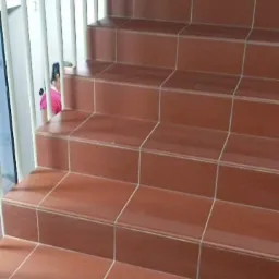 Schody wyłożone terakotą z antypoślizgowymi paskami, biała metalowa balustrada, fragment osoby w różowej bluzce w tle.