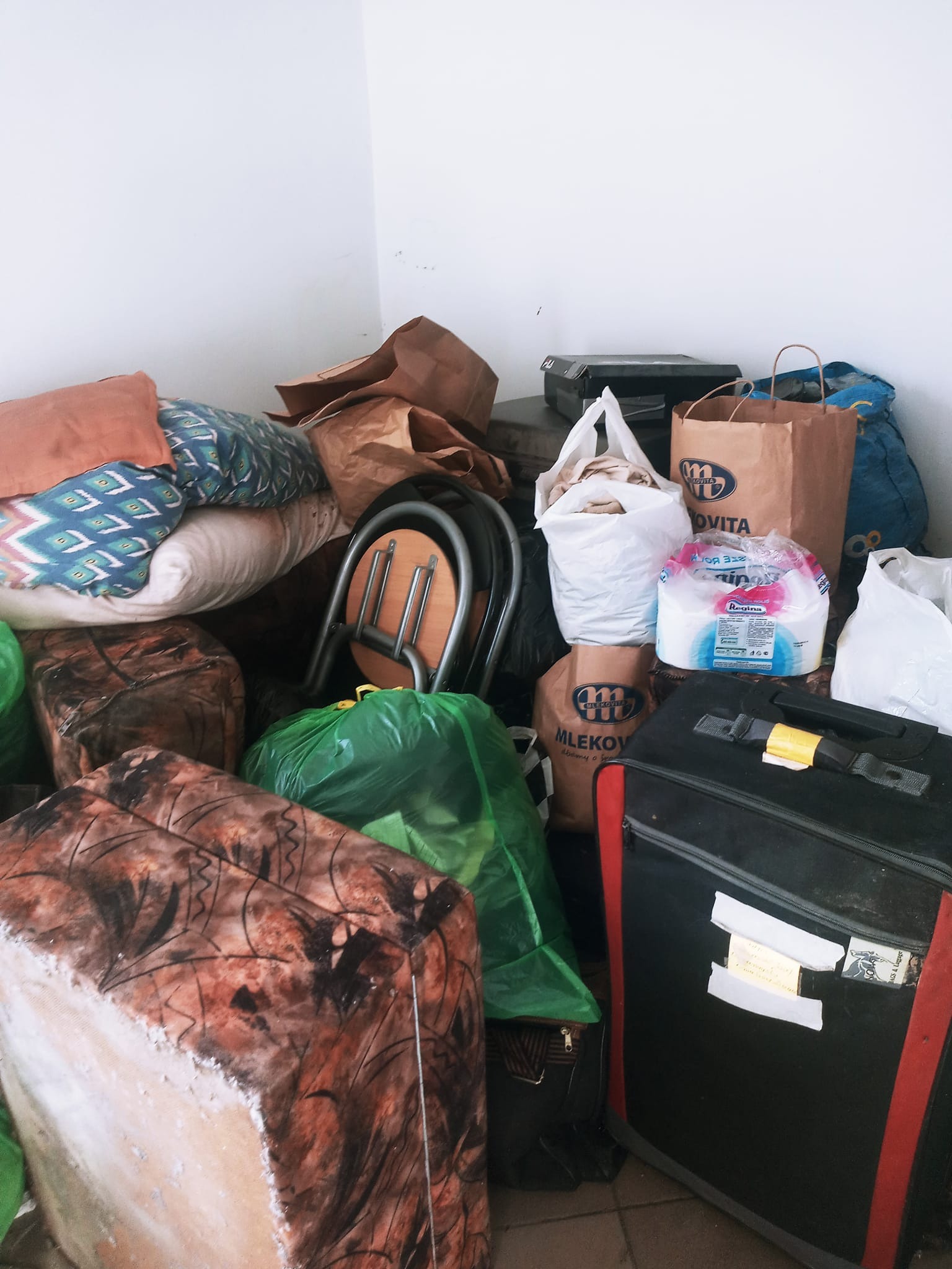 Stos zapakowanych przedmiotów i rzeczy, w tym poduszki, torby papierowe z logo Mlekovita, worki foliowe, walizka z przyklejoną kartką, krzesło, wszystko zgromadzone w rogu pokoju z białymi ścianami.