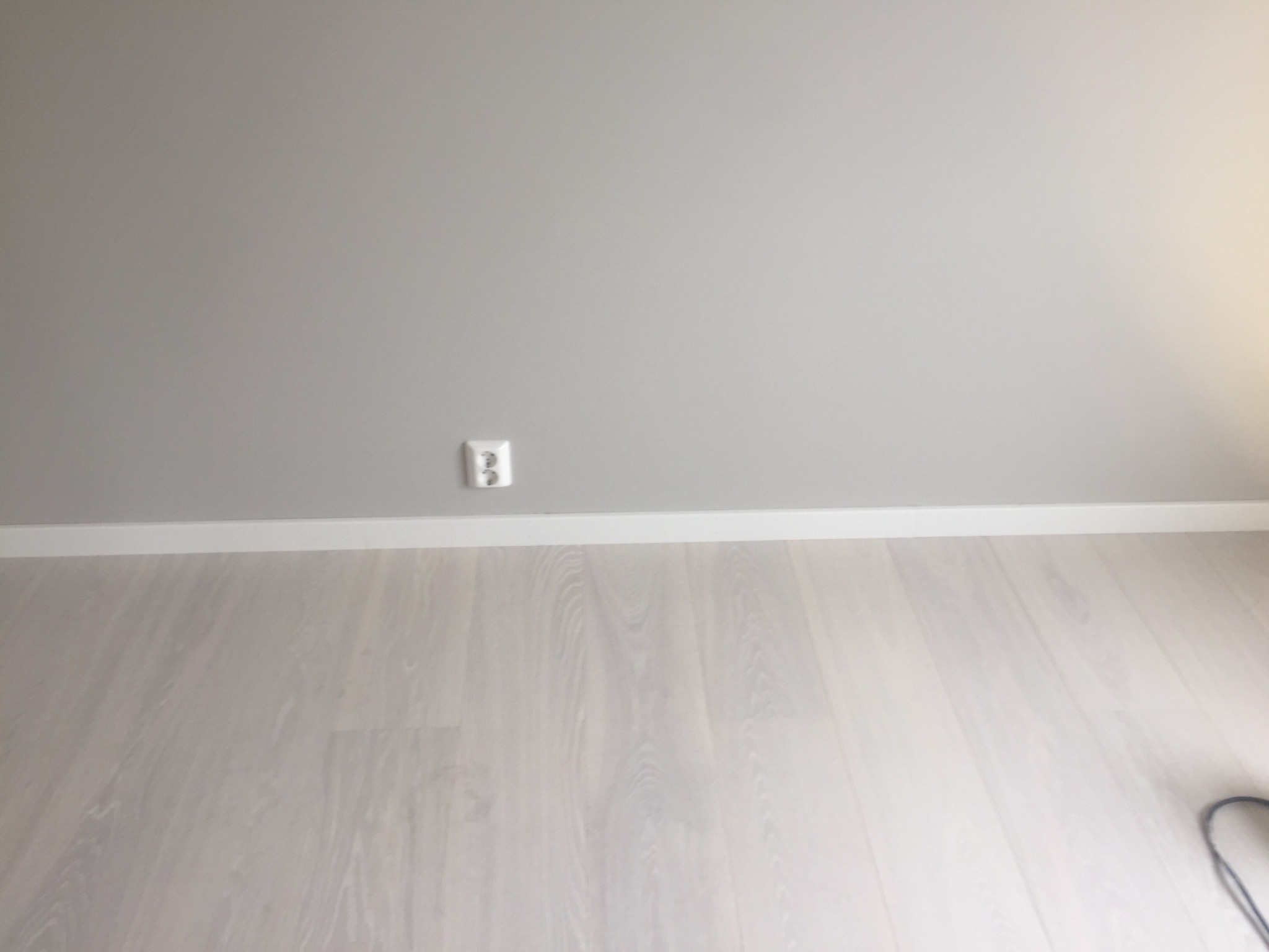 Jasna ściana pomalowana na szaro z gniazdkiem elektrycznym nad białą listwą przypodłogową i podłogą z paneli imitujących jasne drewno.