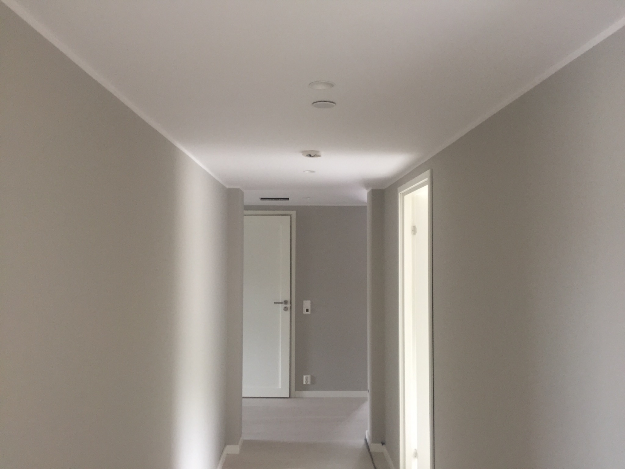 Wnętrze korytarza po remoncie, ściany pomalowane na szaro, biały sufit z wpuszczonymi punktami świetlnymi, widoczne drzwi.