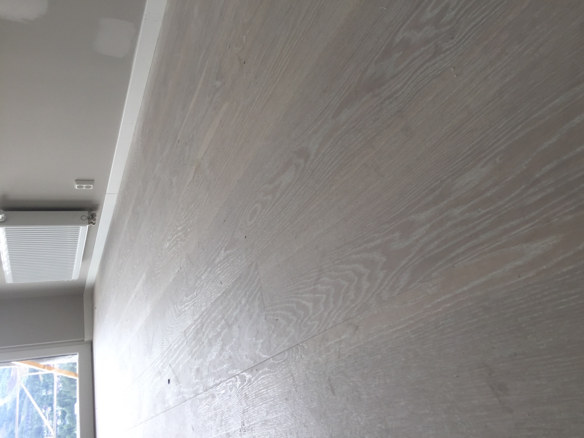 Ściana wykończona panelami drewnianymi o jasnym, bielonym odcieniu, widoczne subtelne słoje drewna, ujęcie z dołu ukazujące fragment sufitu z listwą przypodłogową i grzejnikiem.