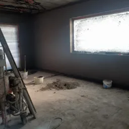 Wnętrze w trakcie prac tynkarskich: ściany pokryte świeżym szarym tynkiem, widoczna maszyna tynkarska, wiaderka i narzędzia na podłodze, okno z zabrudzoną szybą, sufit z widoczną instalacją...