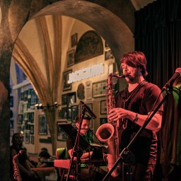 Natalia Mazurkiewicz-Szafarska - Saksofonista na scenie z mikrofonami w klimatycznym wnętrzu. W tle łukowate przejście i ściana z obrazami. Oświetlenie sceniczne.