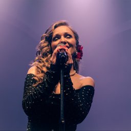 Natalia Mazurkiewicz-Szafarska - Piosenkarka z blond lokami, w czarnej sukience w białe kropki, śpiewa do mikrofonu na tle fioletowej sceny, z czerwoną różą we włosach.