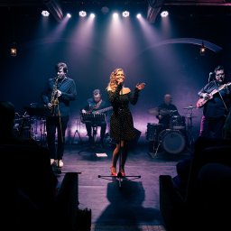 Natalia Mazurkiewicz-Szafarska - Zespół muzyczny na scenie: wokalistka w czarnej sukience, saksofonista, gitarzysta i perkusista. Scena oświetlona reflektorami, widownia w tle.