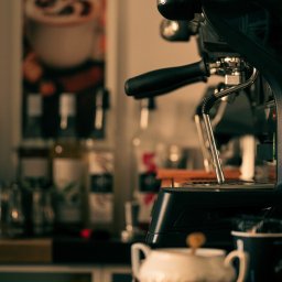 Natalia Mazurkiewicz-Szafarska - Czarny ekspres do kawy w kawiarni, z porcelanową cukiernicą i kubkiem na drewnianym blacie. W tle widoczne butelki i plakat z kawą.