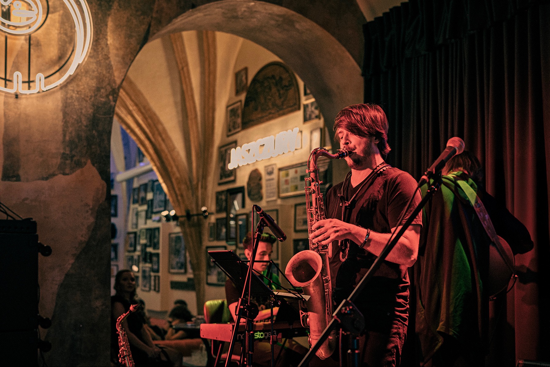 Saksofonista na scenie z mikrofonami w klimatycznym wnętrzu. W tle łukowate przejście i ściana z obrazami. Oświetlenie sceniczne.