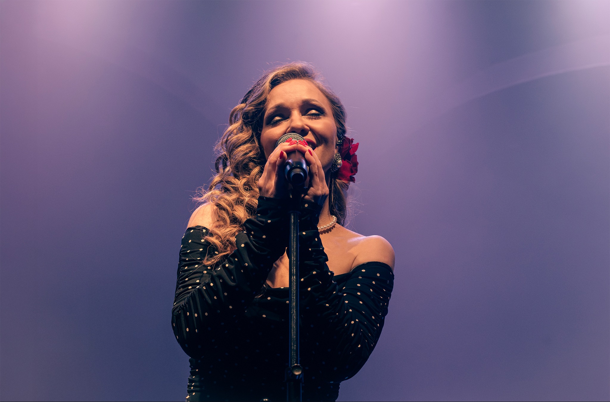 Piosenkarka z blond lokami, w czarnej sukience w białe kropki, śpiewa do mikrofonu na tle fioletowej sceny, z czerwoną różą we włosach.
