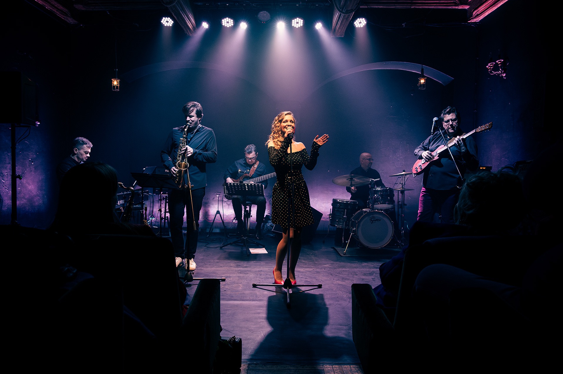 Zespół muzyczny na scenie: wokalistka w czarnej sukience, saksofonista, gitarzysta i perkusista. Scena oświetlona reflektorami, widownia w tle.