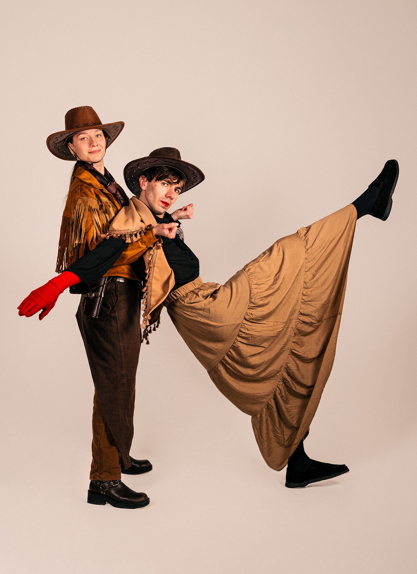 Dwie osoby w stylizowanych strojach kowbojskich pozują na jasnym tle. Jedna osoba stoi, druga w dynamicznej pozie z uniesioną nogą. Nietypowy, artystyczny portret.