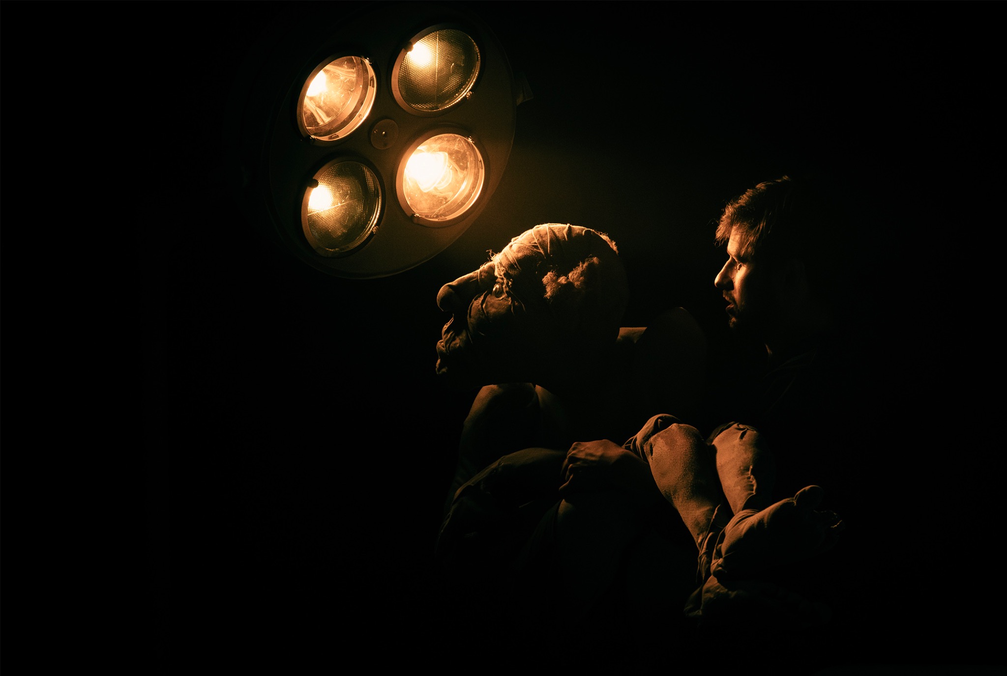 Mroczna fotografia studyjna: mężczyzna w profilu, oświetlony lampą chirurgiczną, obok rzeźby głowy z bandażami. Klimatyczne, teatralne oświetlenie.