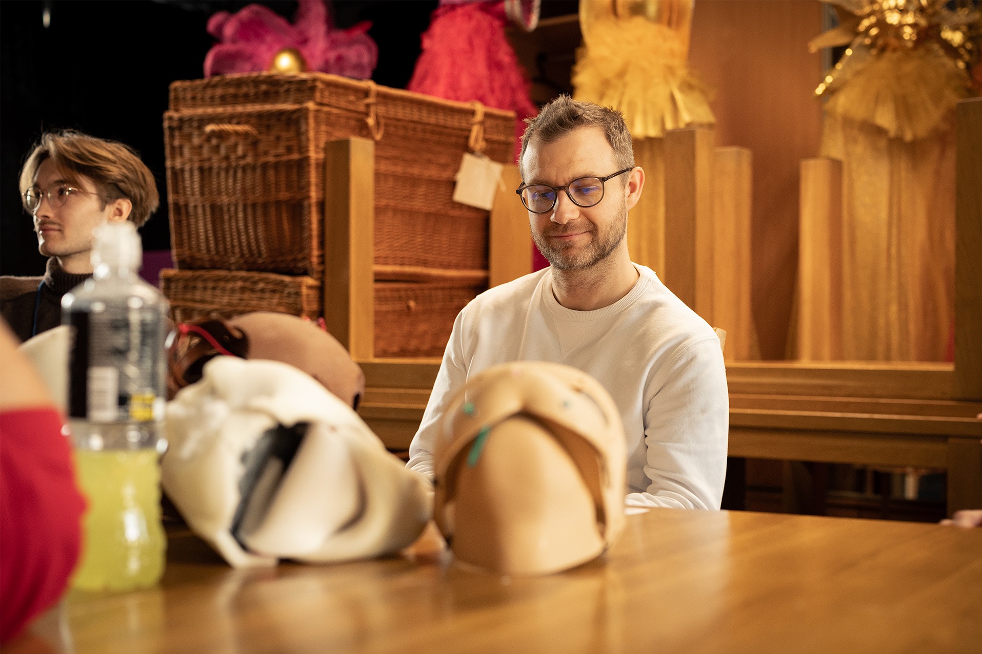 Mężczyzna w okularach siedzi przy stole z manekinami głów, otoczony dekoracjami, w tle widoczny drugi mężczyzna. Zdjęcie z bliska, ciepłe oświetlenie.