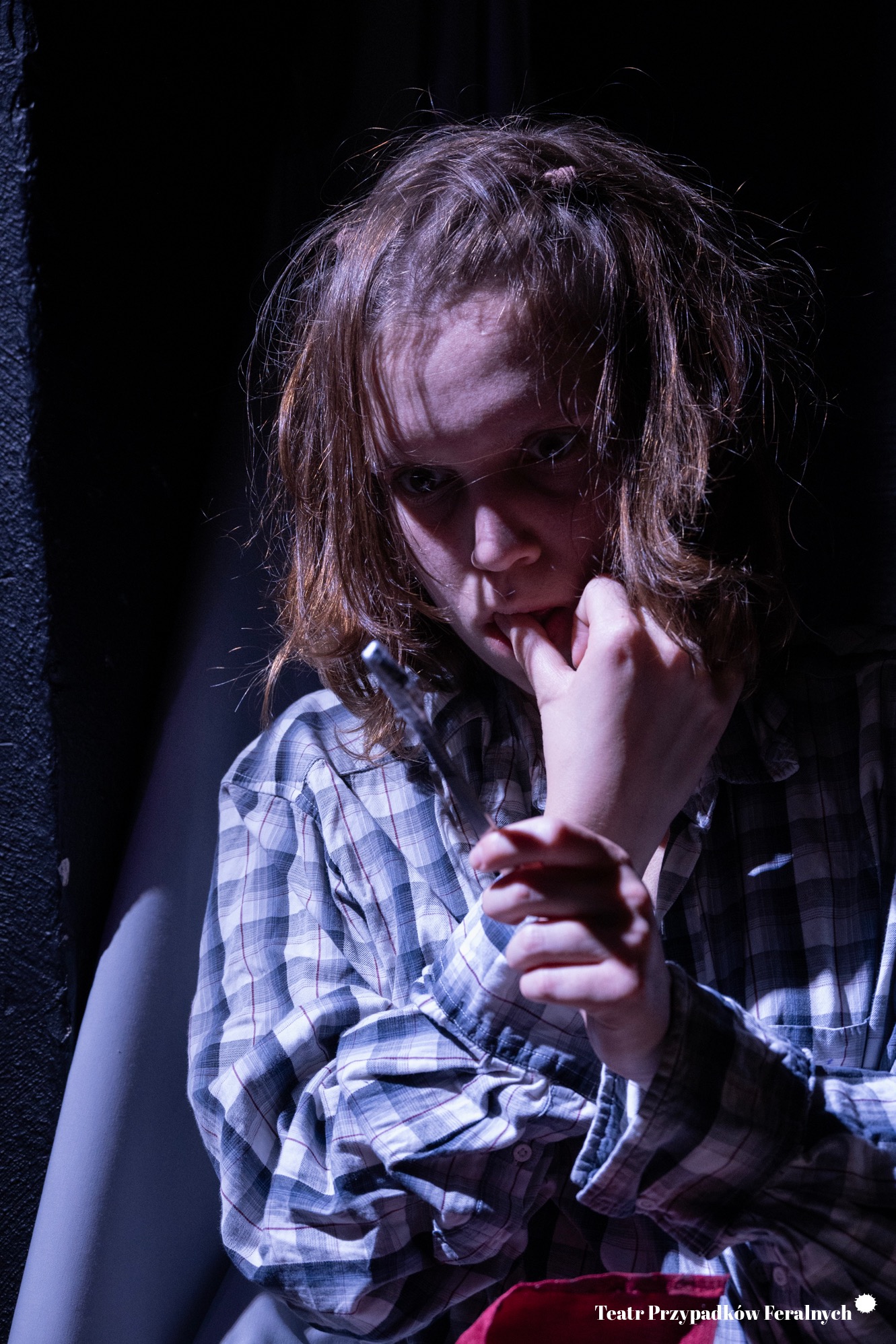 Portret młodej osoby w kraciastej koszuli, w mrocznym oświetleniu, z ręką przy ustach, w teatralnej scenerii. Ujęcie z dołu, widoczne logo teatru.
