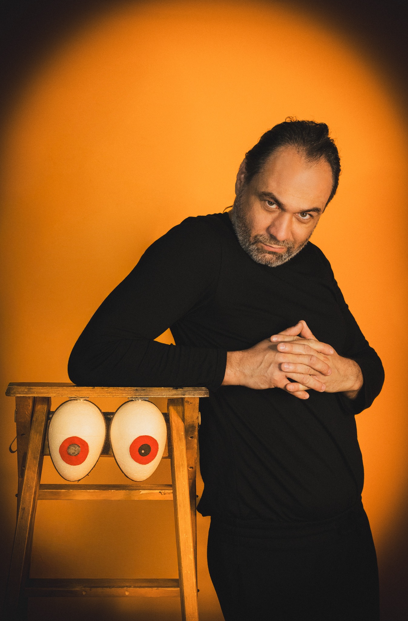 Mężczyzna w czerni opiera się o drewnianą drabinę z dwoma dużymi, stylizowanymi oczami na pomarańczowym tle, patrząc wprost w obiektyw z lekkim uśmiechem.