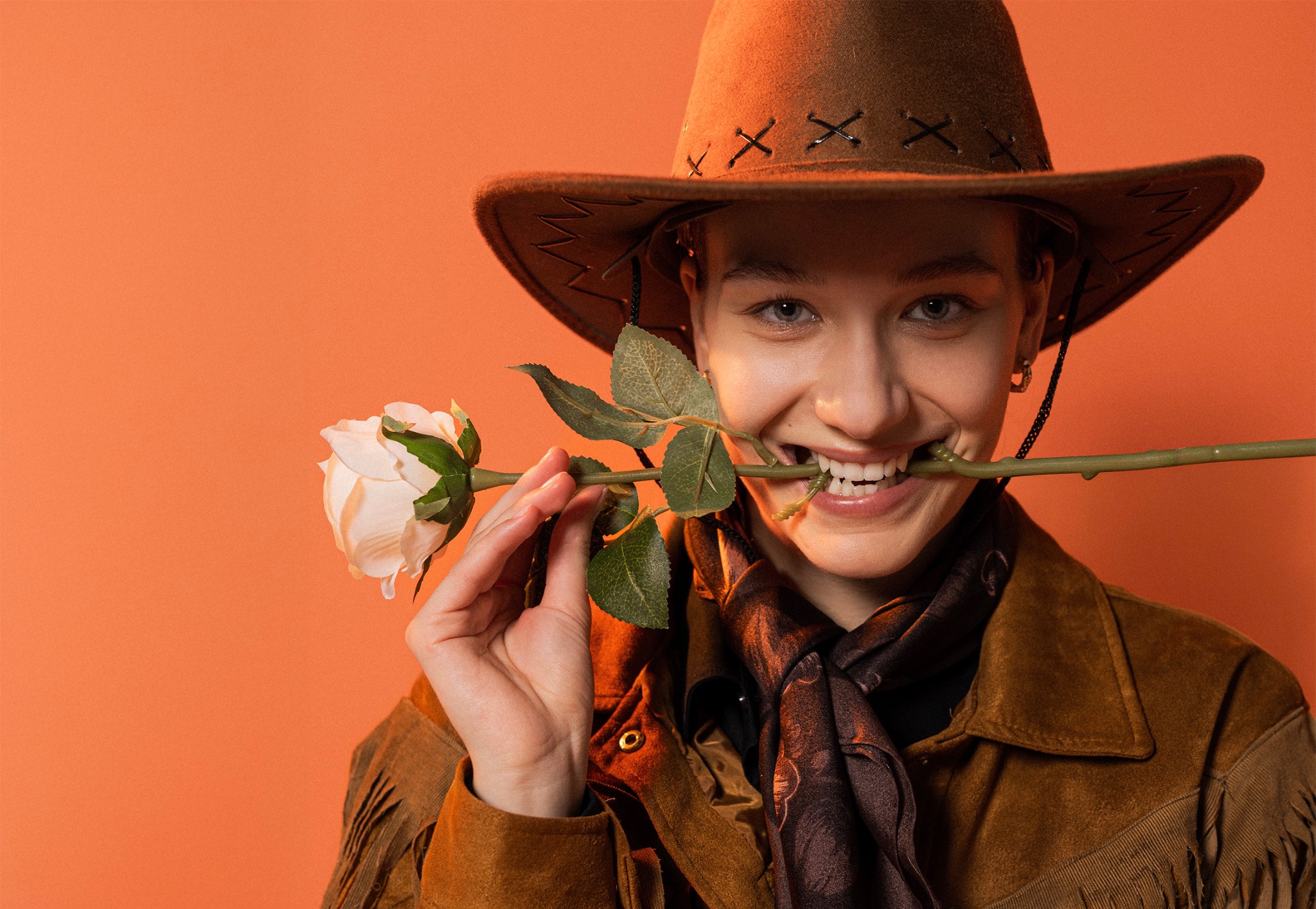 Uśmiechnięta kobieta w kapeluszu kowbojskim, trzymająca białą różę w ustach, na pomarańczowym tle. Ubrana w zamszową kurtkę z frędzlami i wzorzysty szal.