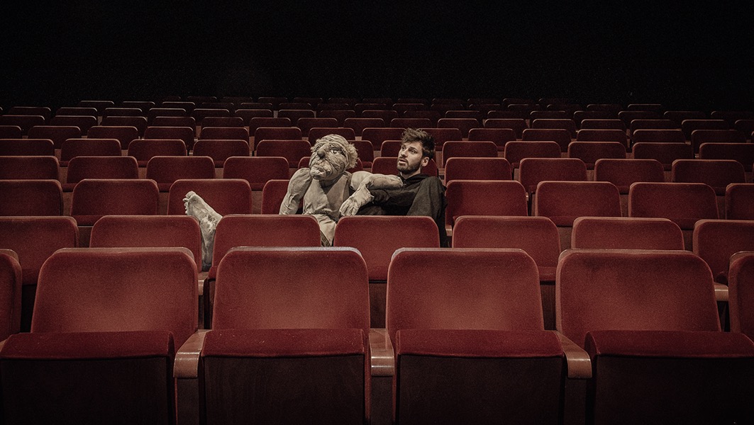 Mężczyzna z brodą siedzi w kinie obok kukiełki, która wystawia stopę na siedzenie. Rzędy czerwonych foteli wypełniają kadr, a czarne tło dodaje teatralności.
