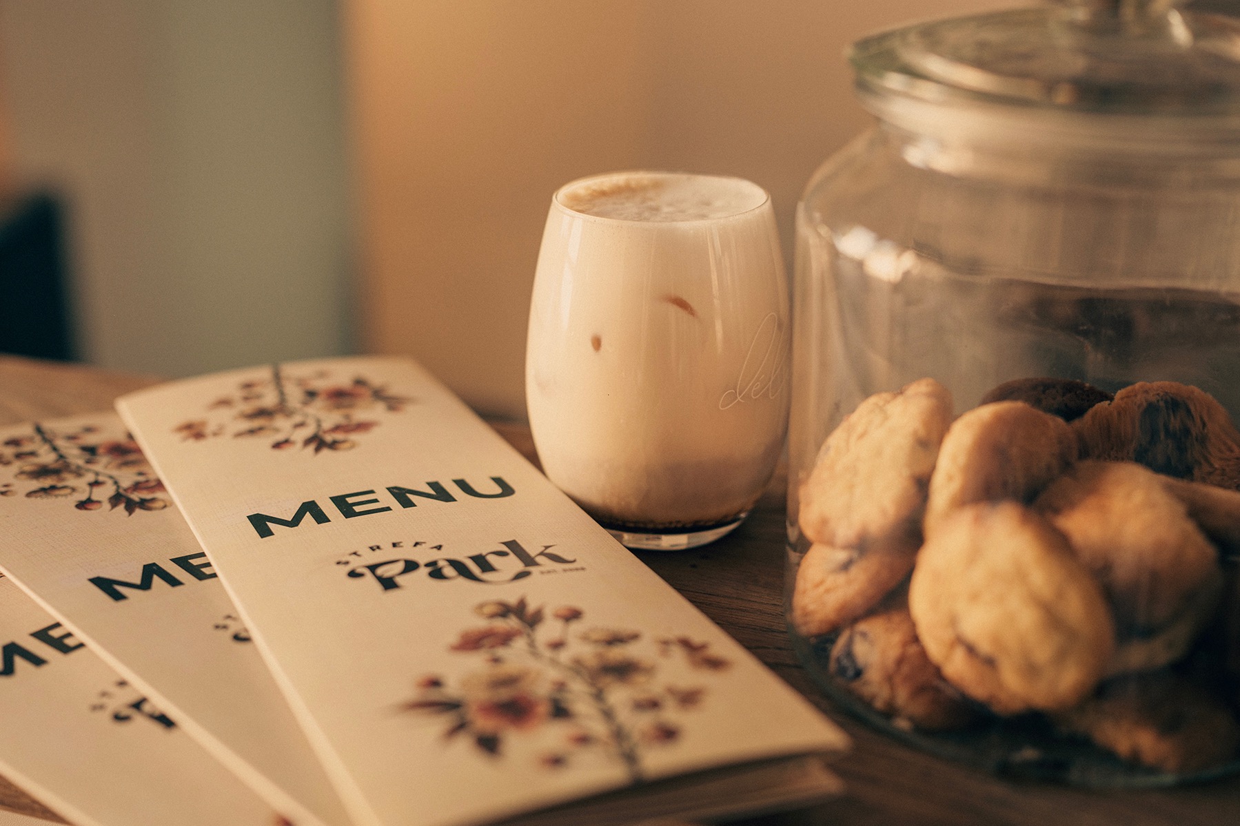 Stylowe menu 'Strefa Park' z kwiatowym motywem, obok szklanka latte i słoik ciastek. Ciepłe, stonowane barwy, delikatne oświetlenie, przytulna atmosfera kawiarni.