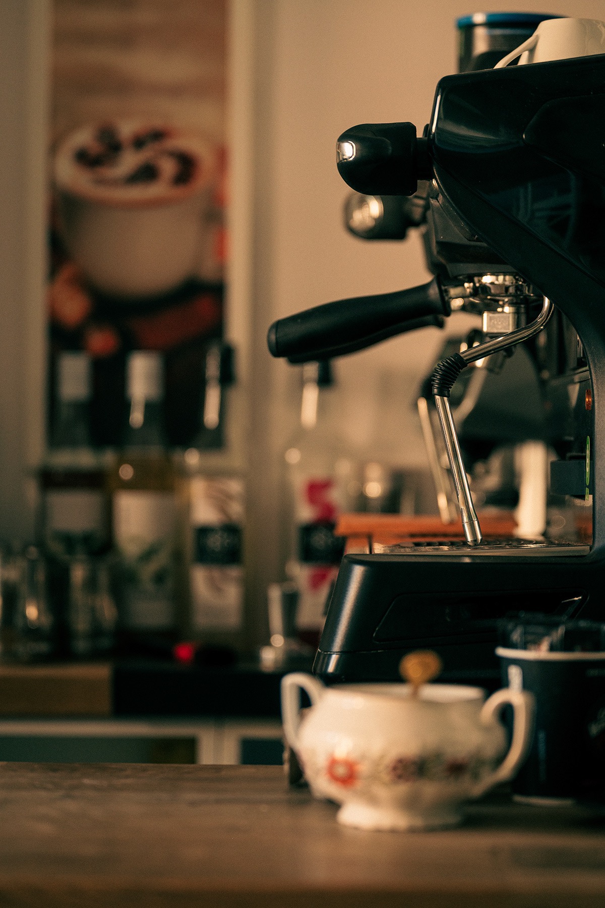 Czarny ekspres do kawy w kawiarni, z porcelanową cukiernicą i kubkiem na drewnianym blacie. W tle widoczne butelki i plakat z kawą.