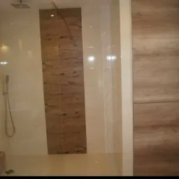 Nowoczesna kabina prysznicowa z deszczownicą i panelami imitującymi drewno, połączona z zabudową meblową o podobnym wzorze, tworząca spójną aranżację.