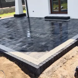 Świeżo ułożona, mokra, grafitowa kostka brukowa na tarasie z obrzeżem z jasnego grysu i czarnych krawężników. Widoczna zagęszczarka wibracyjna i paleta z kostką w tle.