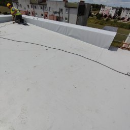 Dach-bud Dariusz matecki - Pracownik w żółtym kasku i kamizelce odblaskowej przy pracy na płaskim, jasnym dachu, prawdopodobnie podczas montażu izolacji, z widocznymi budynkami mieszkalnymi w tle.