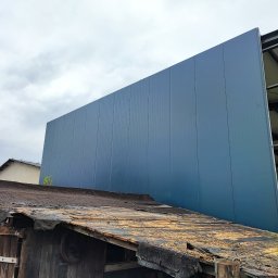 Dach-bud Dariusz matecki - Zniszczony dach drewnianej konstrukcji na tle nowoczesnej, niebieskiej hali przemysłowej z blachy trapezowej.