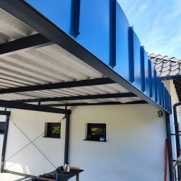 Dach-bud Dariusz matecki - Nowoczesna wiata garażowa z niebieskimi panelami blaszanej obróbki, czarną konstrukcją stalową i widocznym podsufitką z płyt, narzędzia na stole w tle.