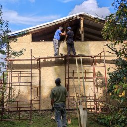 Dach-bud Dariusz matecki - Dwóch dekarzy na rusztowaniu montuje płytę OSB na dachu budynku, trzeci pracownik obserwuje z dołu, widoczne drzewa i niebo z chmurami w tle.