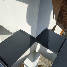 Dach-bud Dariusz matecki - Szczegółowe ujęcie połączenia elementów blaszanych na dachu w Jarocinie, widoczny fragment białej ściany i podłoże z gruzem, rzucające cień na konstrukcję.