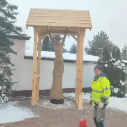 Pracownik w odblaskowej kurtce stoi obok drewnianej konstrukcji zadaszenia nad rzeźbą na zewnątrz w zimowy dzień.