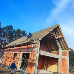 Dach-bud Dariusz matecki - Budowa domu jednorodzinnego w trakcie, widoczne rusztowanie, niezadaszona konstrukcja dachu z łatami, murarz na dachu, drugi pracownik na ziemi, cegły i betonowe elementy konstrukcyjne.