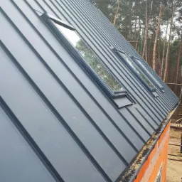 Nowy, grafitowy dach z blachy na surowej, ceglanej konstrukcji domu, z trzema oknami dachowymi, na tle lasu.