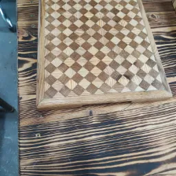 Drewniany blat stołu z wzorem szachownicy, wykonany z jasnych i ciemnych kwadratów drewna, na tle blatu z wypalanymi słojami.