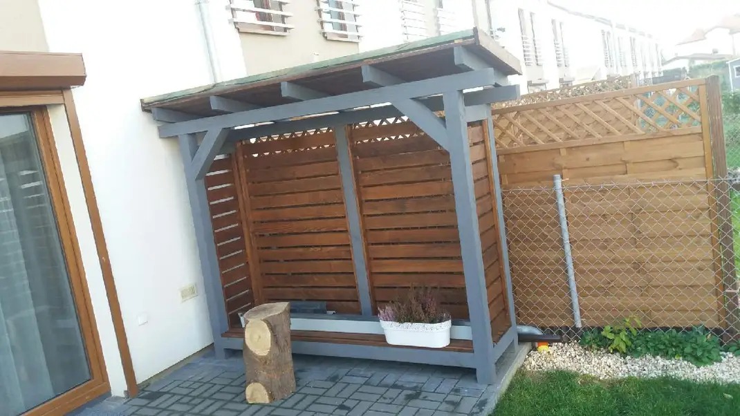 Drewniana altana przy ścianie budynku z siedziskiem, dekoracją z kwiatów w doniczce i pniakiem, tło stanowi płot z siatki i drewniane panele.