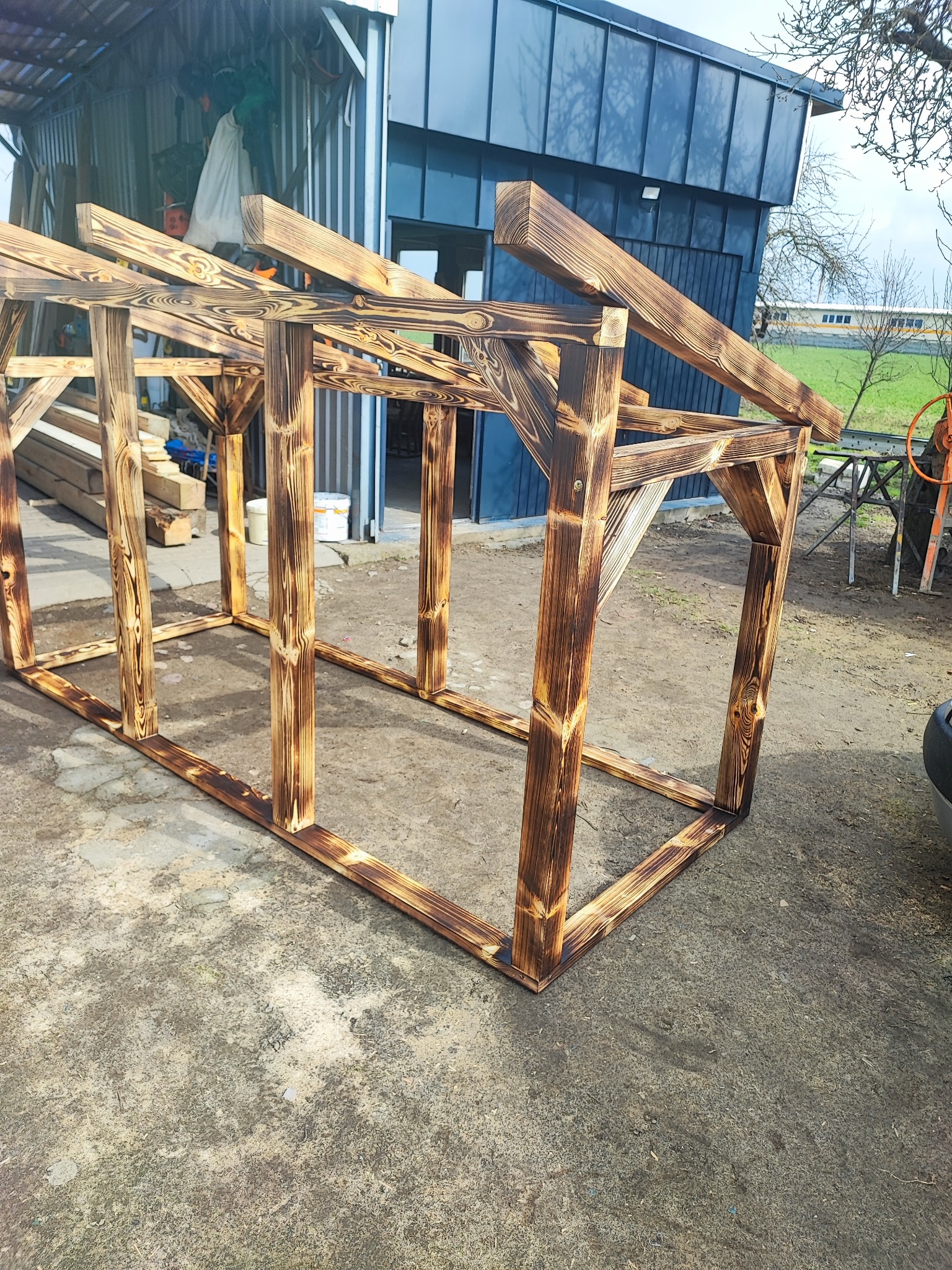 Szkielet drewnianej konstrukcji z belek opalanych ogniem, przygotowany do budowy, widoczny na zewnątrz budynku gospodarczego.