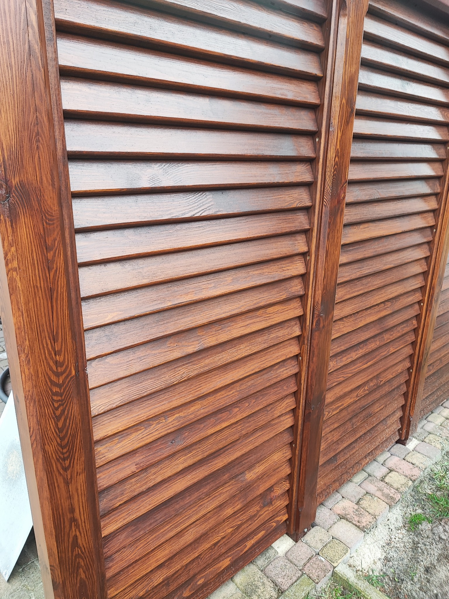 Drewniana konstrukcja z żaluzjami w kolorze ciemnego brązu, widoczna z boku na tle kostki brukowej i trawy.