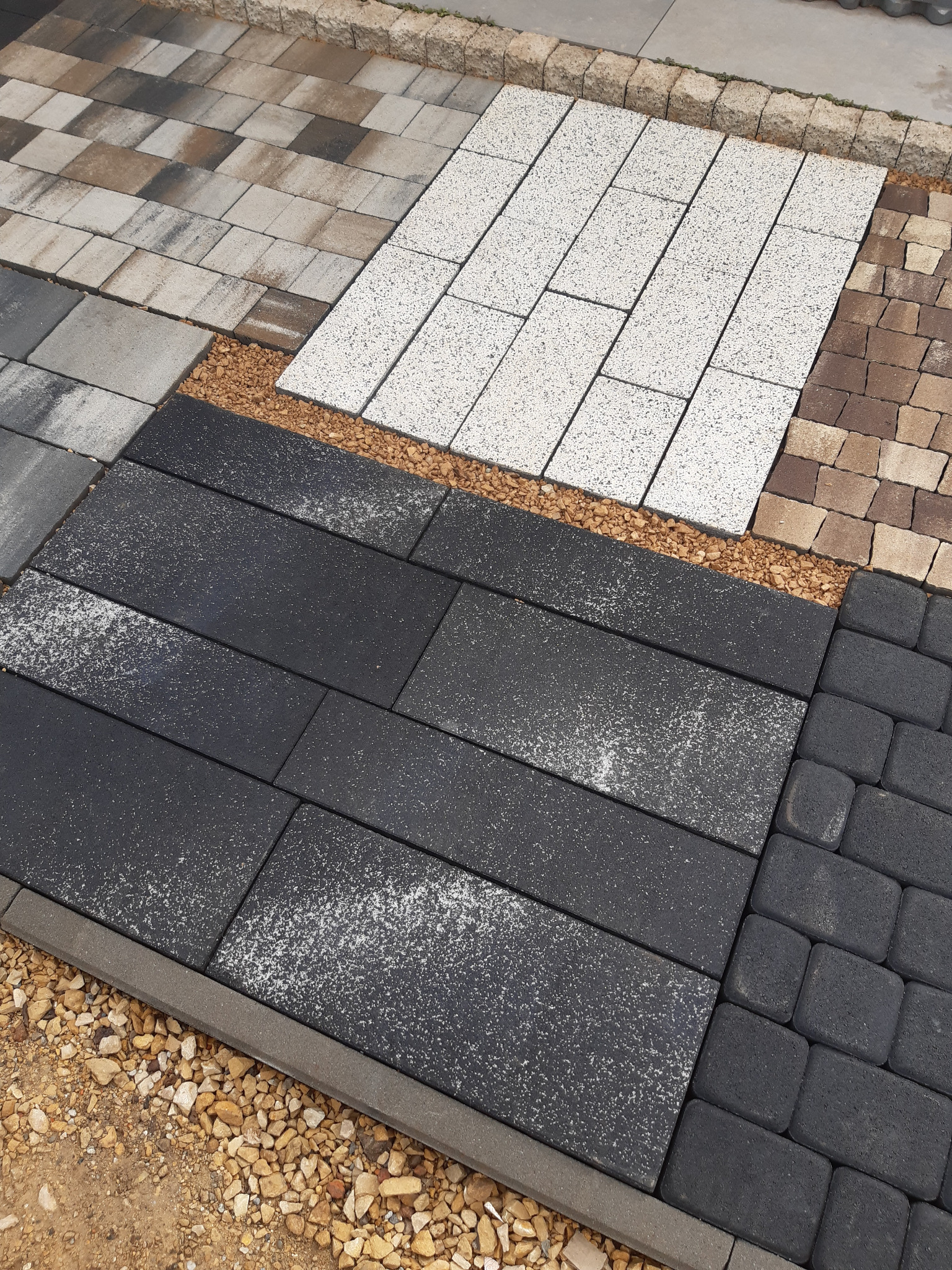 Ekspozycja różnych wzorów i kolorów nawierzchni z kostki brukowej, oddzielonych drobnym żwirem, prezentowanych na placu budowy.