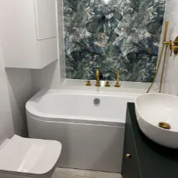 Nowoczesna łazienka z umywalką nablatową, wanną i toaletą w zabudowie, ozdobiona płytkami z motywem roślinnym i złotą armaturą.