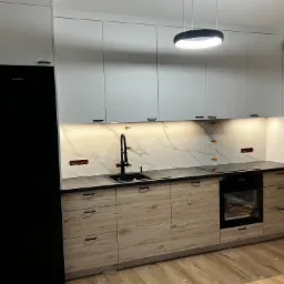 Nowoczesna kuchnia z drewnianymi szafkami dolnymi i białymi górnymi, czarny zlew i płyta indukcyjna, marmurowy backsplash z widocznymi otworami na gniazdka i podświetleniem LED, czarna lodówka...